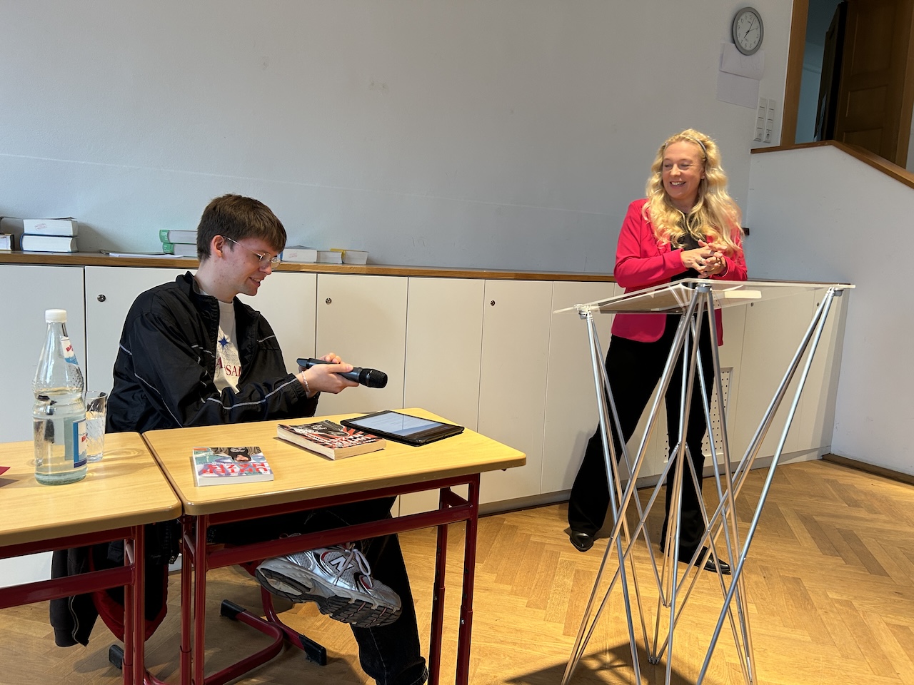 Frau am Rednerpult, junger Mann sitzend am Tisch