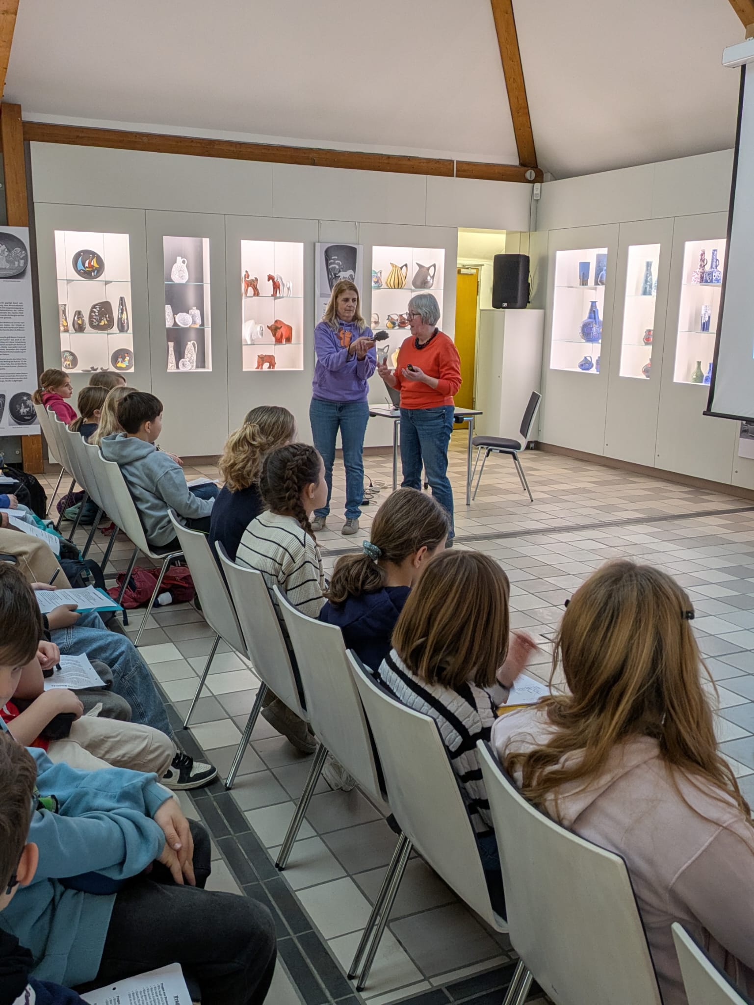 Redakteurin hält einer Dame ein Mikro hin, Schülerinnen und Schüler sitzen in riehen davor, im Hintergrund Kunstwerke aus Glas in Vitrinen