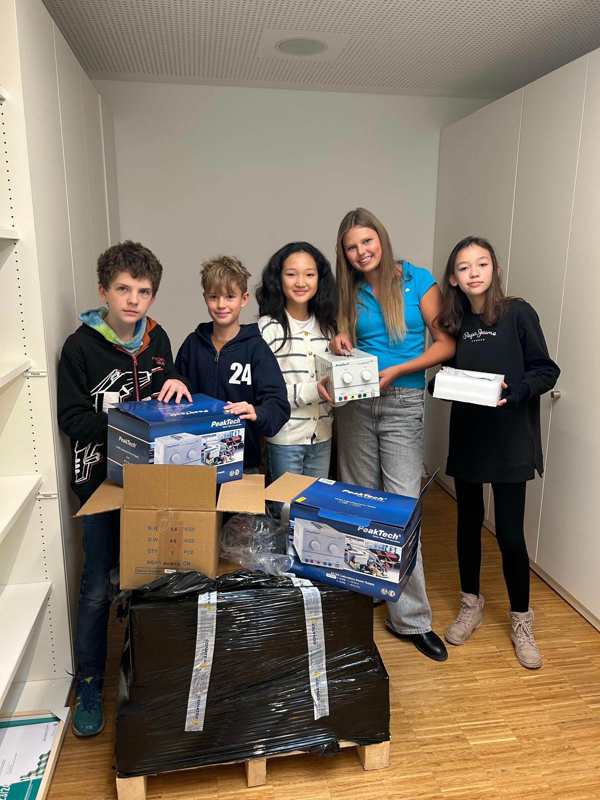 Zwei Jungen und drei Mädchen hinter einem großen Packet, Wechselstromgeräte in der Hand