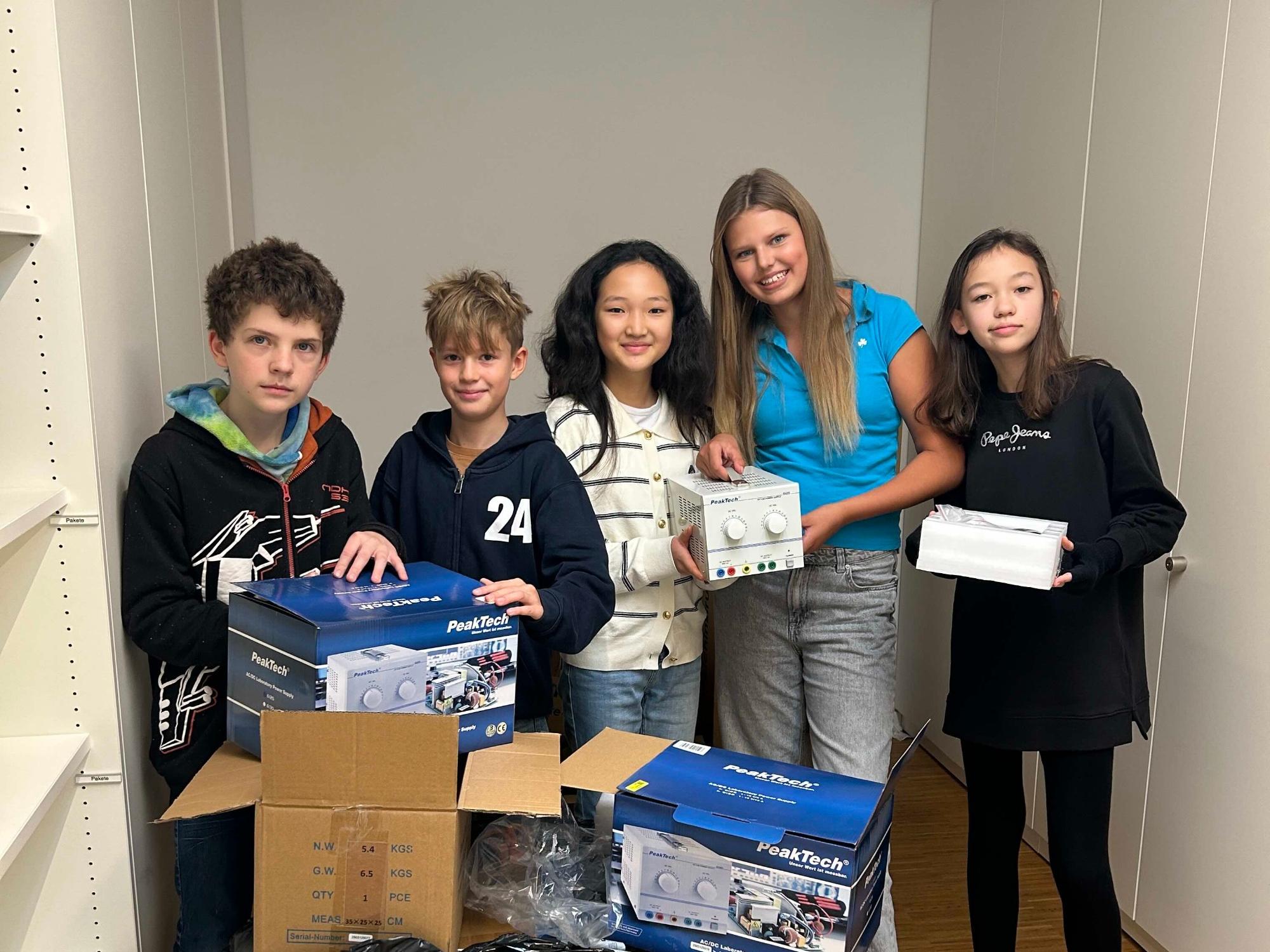 Zwei Jungen und drei Mädchen hinter einem großen Packet, Wechselstromgeräte in der Hand