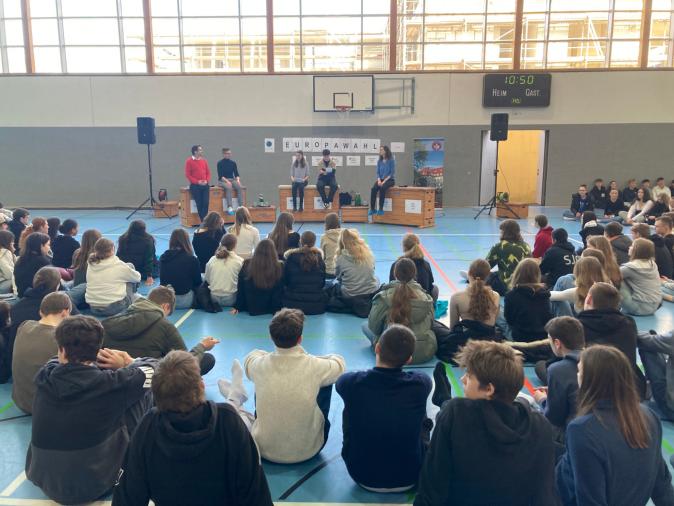 Schülerinnen und Schüler sitzen auf dem Boden der Turnhalle mit Blick auf die Politiker:innen und Moderator:innen