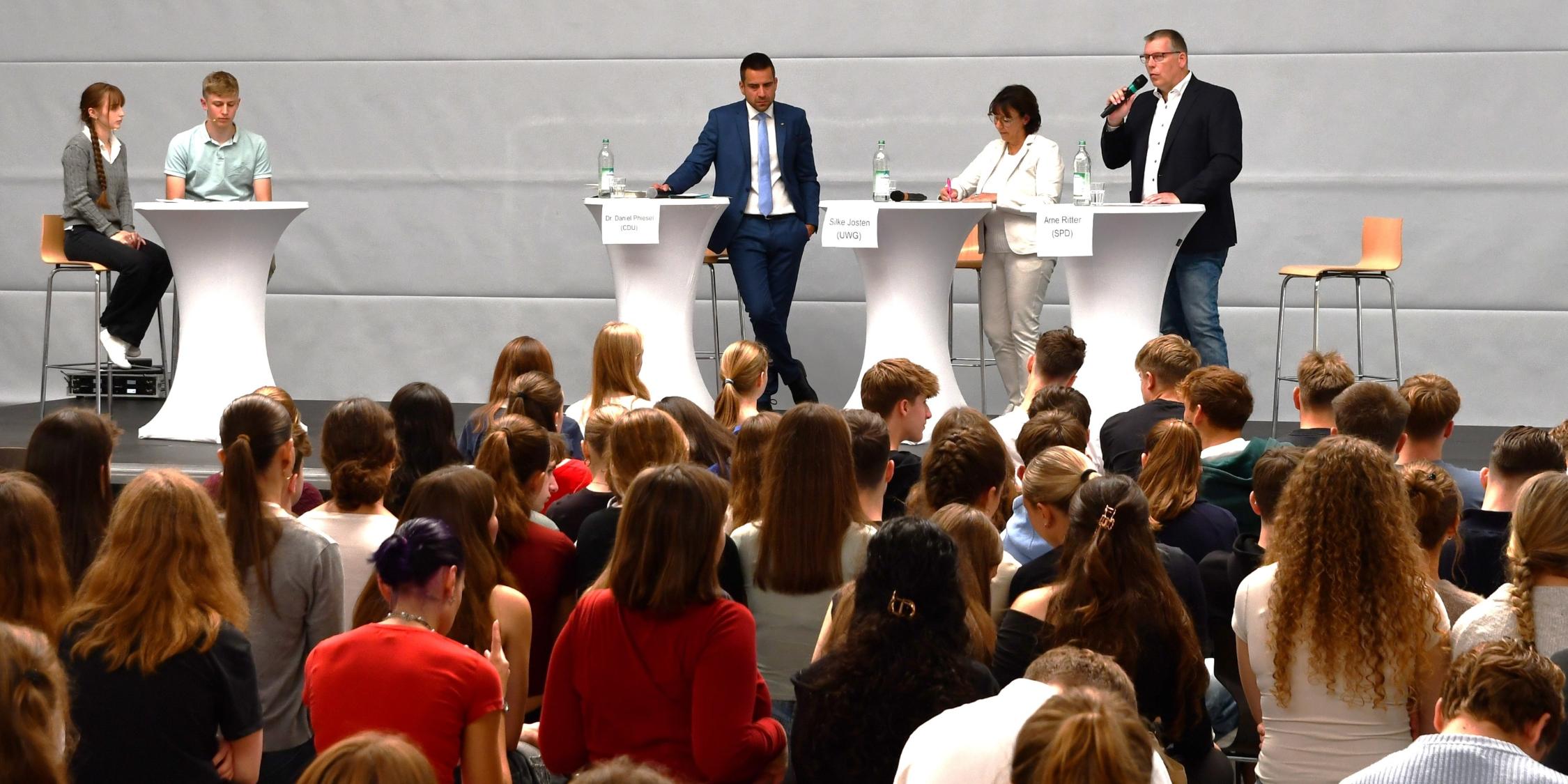 zwei Männer, eine Frau an Stehtischen, einer mit Mikro. Ein weiterer Stehtisch mit einer Schülerin und einem Schüler, Jugendliche