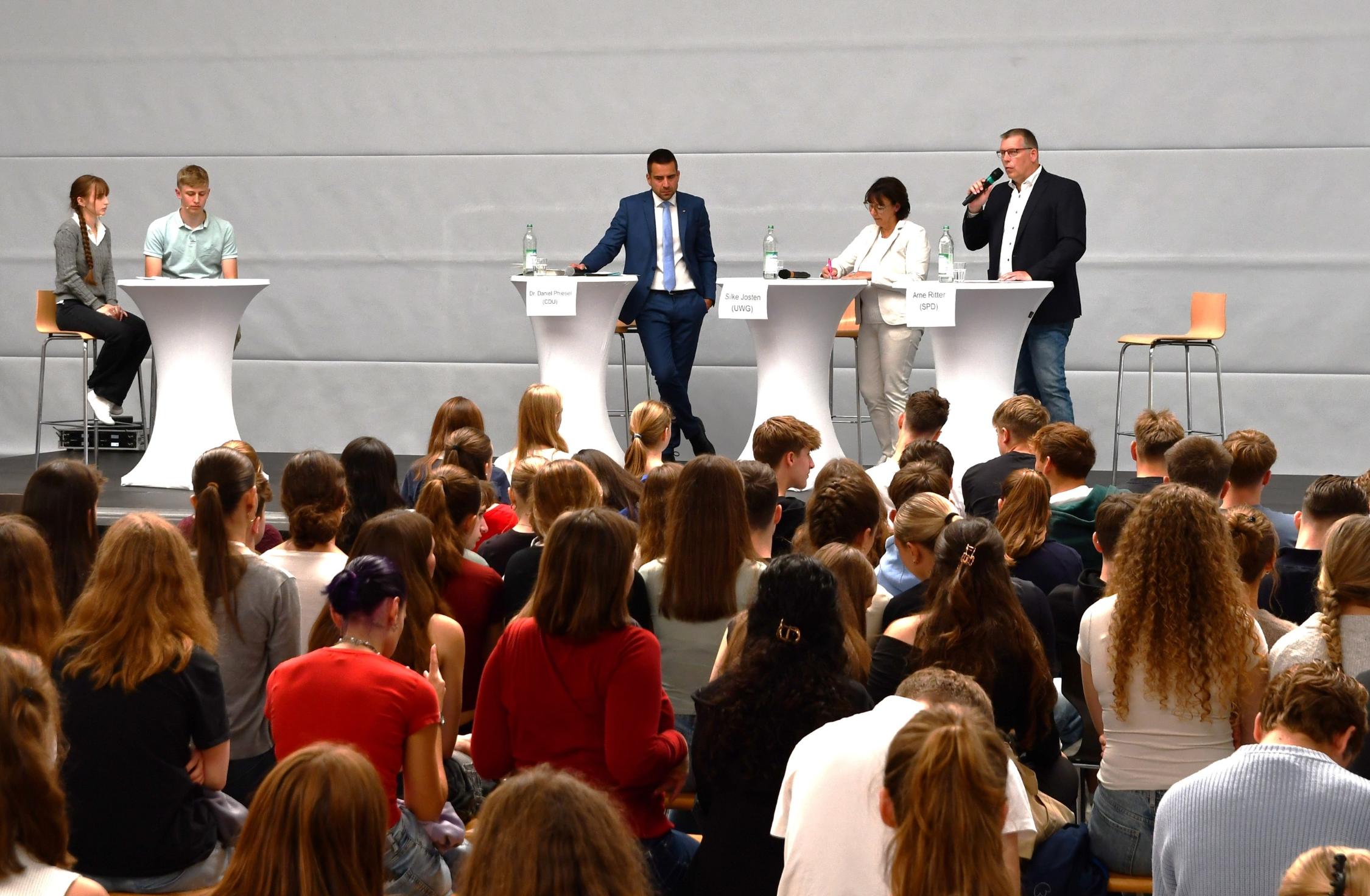 zwei Männer, eine Frau an Stehtischen, einer mit Mikro. Ein weiterer Stehtisch mit einer Schülerin und einem Schüler, Jugendliche