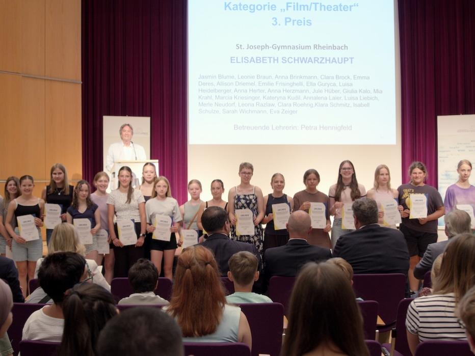 Mädchengruppe mit Urkunden in der Hand auf einer Bühne, davor Publikum sitzend