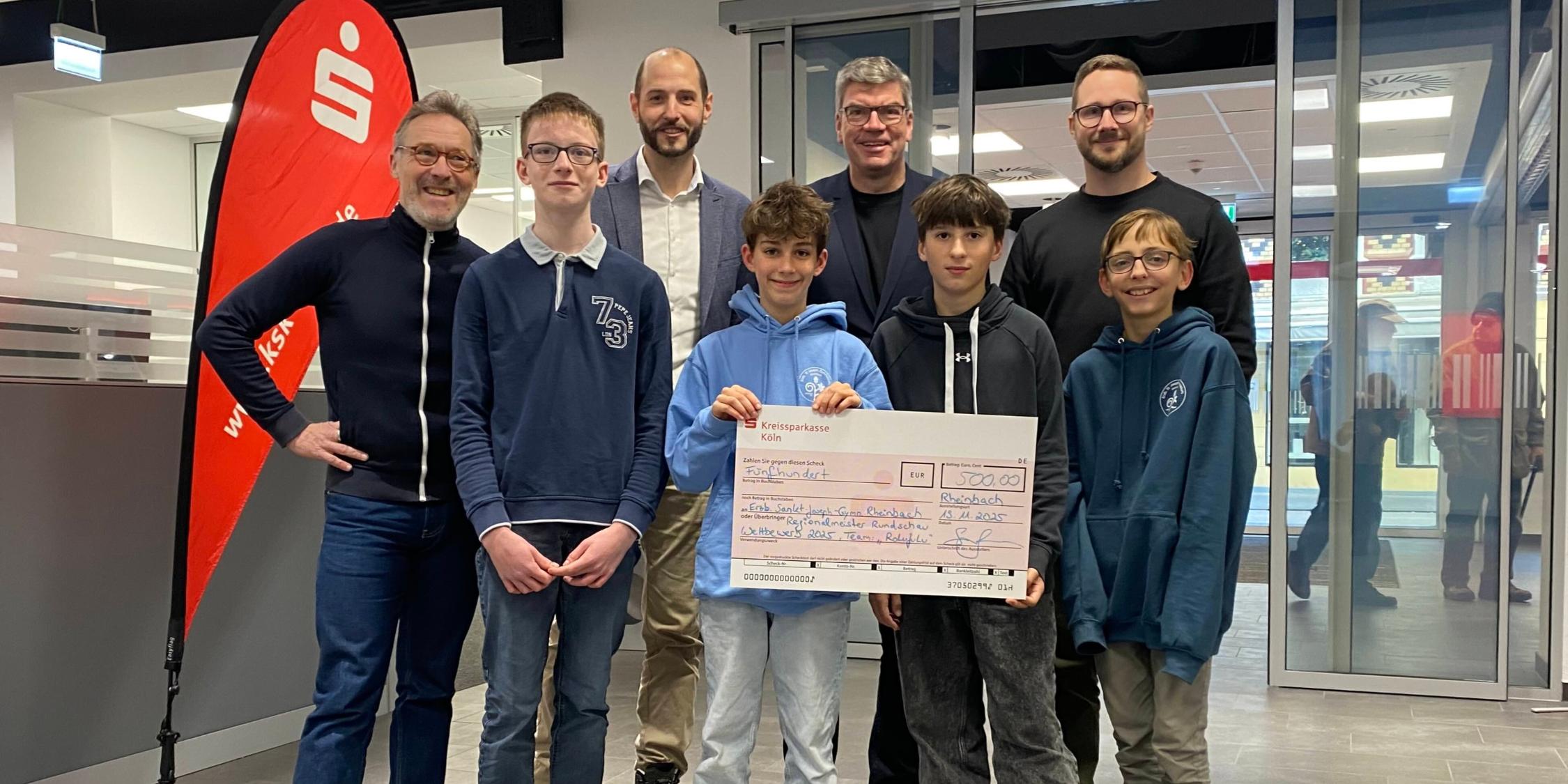 vier Jungen, einer hält einen übergroßen Scheck mit dem Betrag 500 Euro, in der zweiten Reihe vier Männer