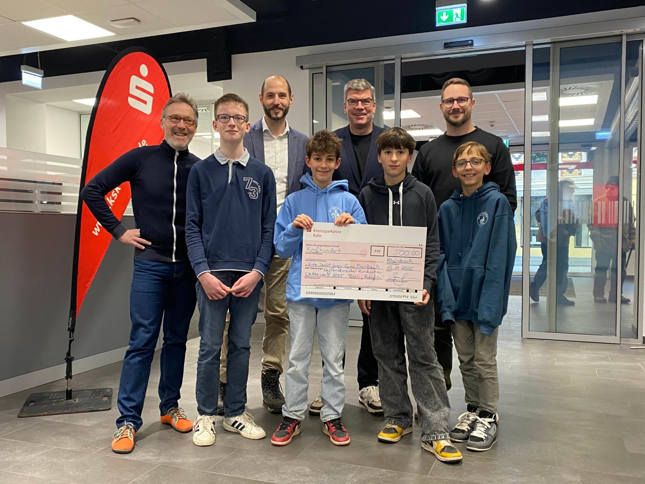 vier Jungen, einer hält einen übergroßen Scheck mit dem Betrag 500 Euro, in der zweiten Reihe vier Männer