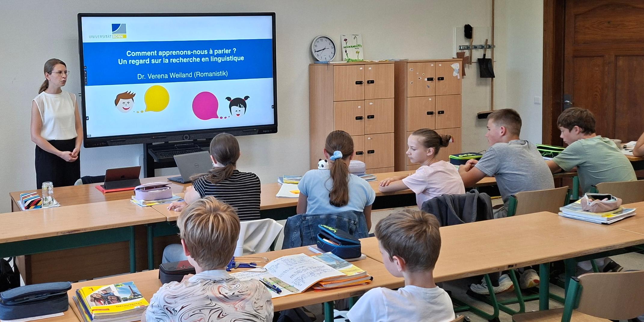 Frau steht vor einem digitalen Board, Schüler:innen sitzen in Reihen in der Klasse