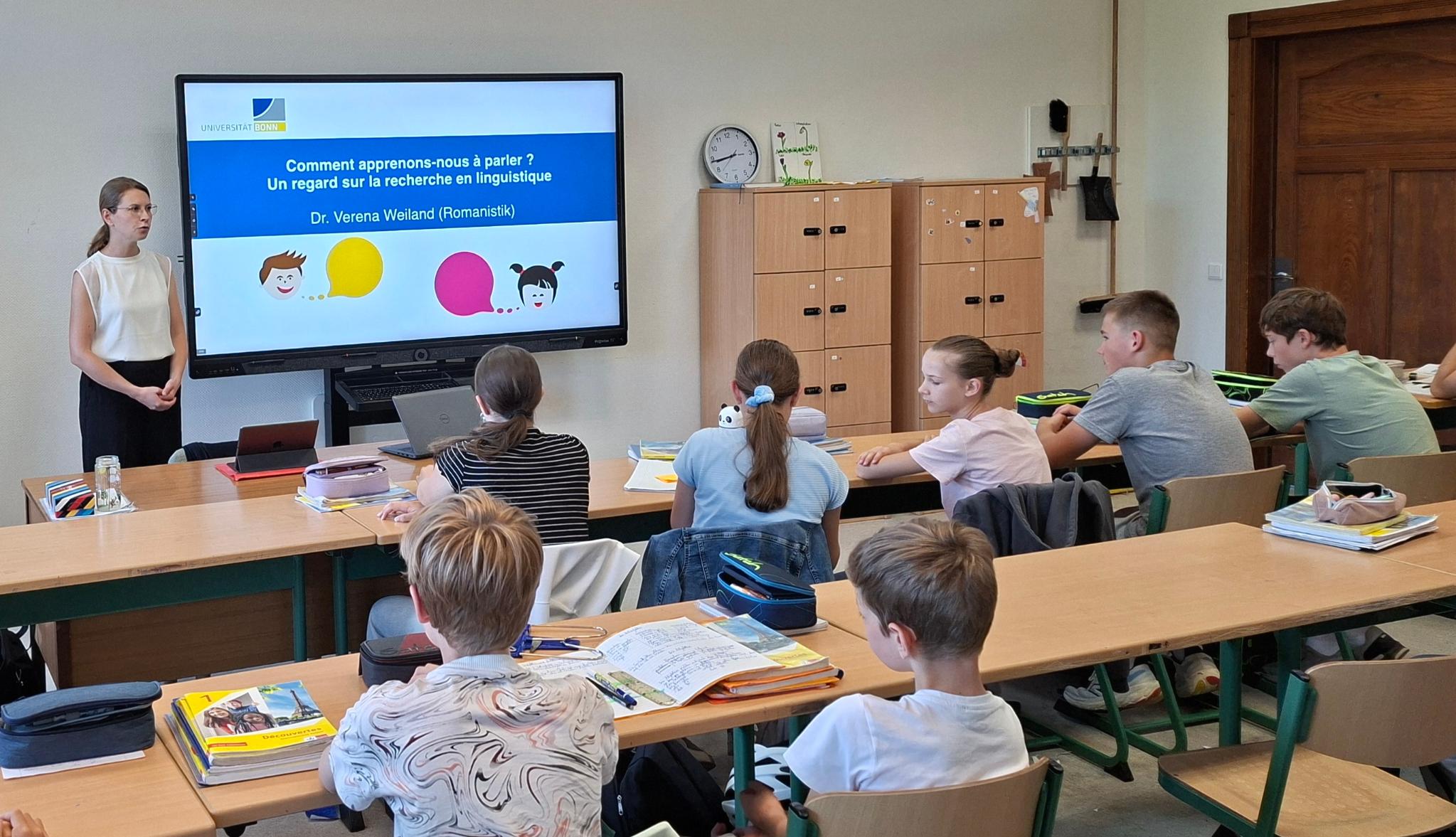 Frau steht vor einem digitalen Board, Schüler:innen sitzen in Reihen in der Klasse
