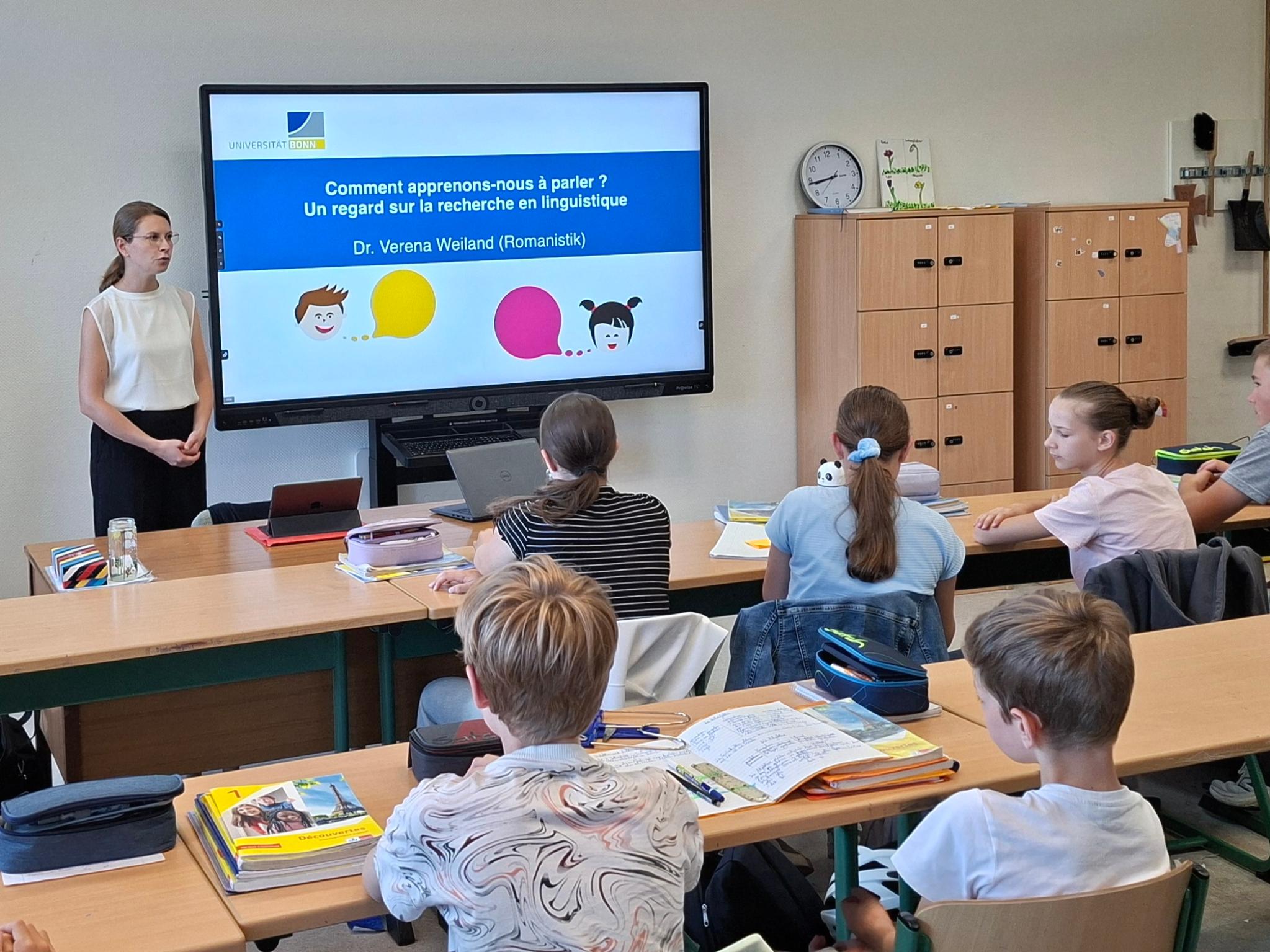 Frau steht vor einem digitalen Board, Schüler:innen sitzen in Reihen in der Klasse