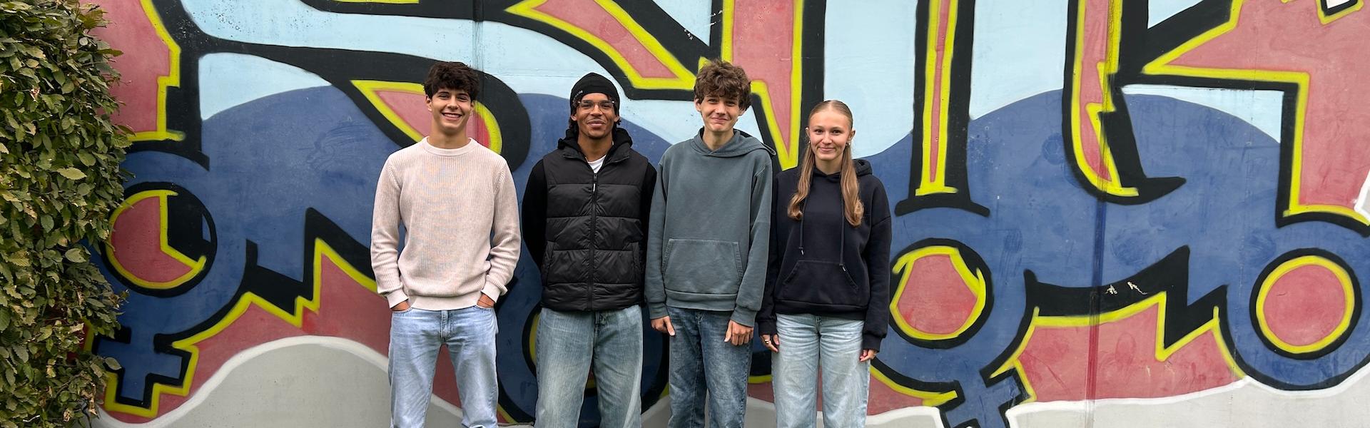 drei Jungen, ein Mädchen vor einem Graffiti mit den Buchstaben SJG