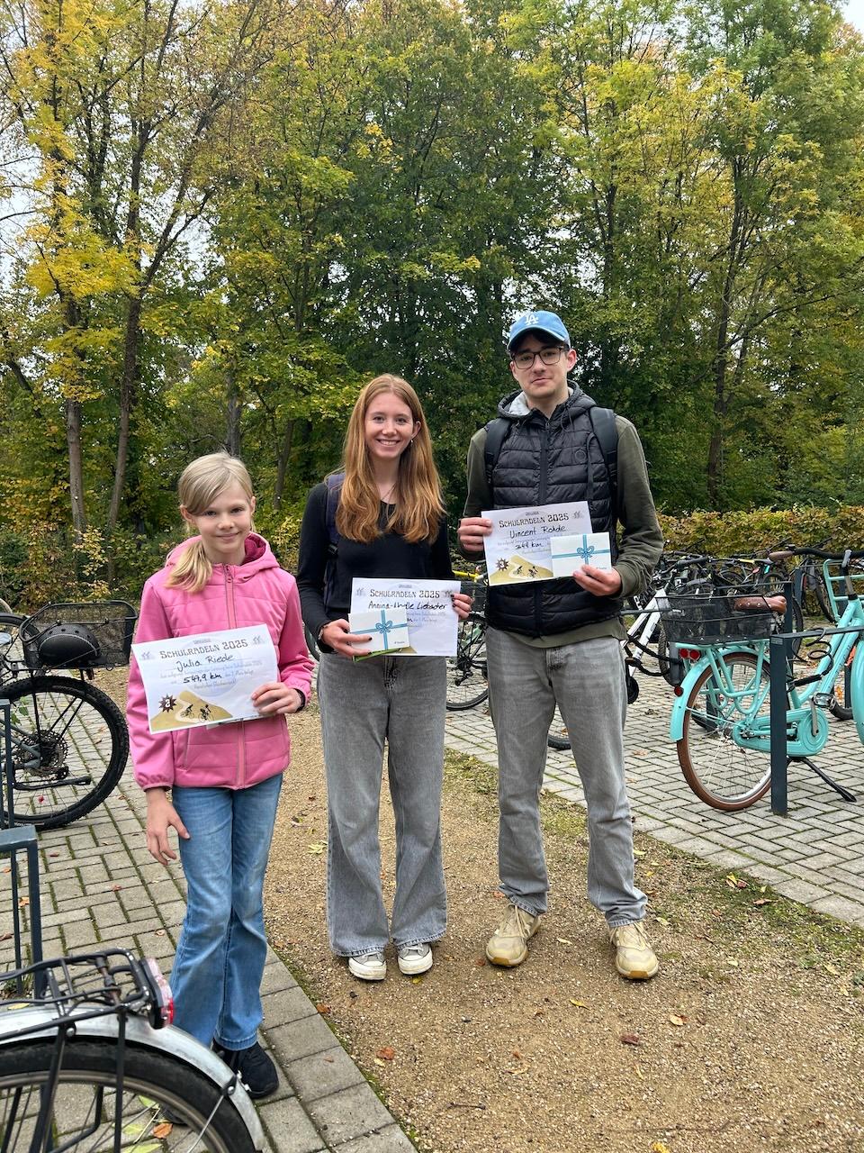 ein Kind, eine Jugendliche, ein Jugendlicher mit Urkunden in der Hand, daneben Fahrräder