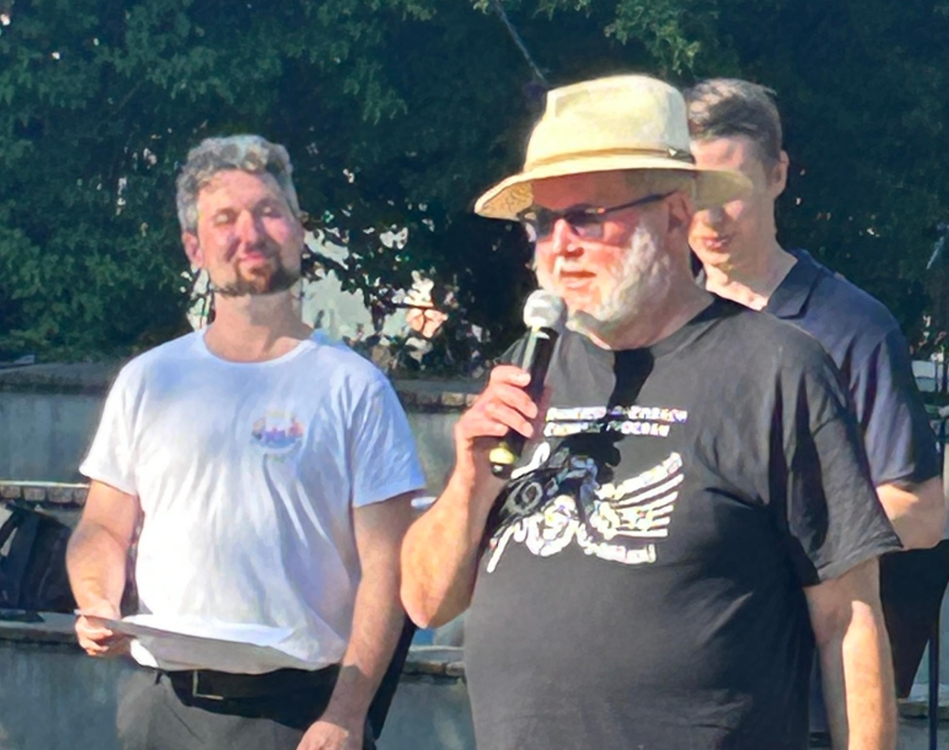 drei Männer stehend, ein Mann mit Hut mit Mikro in der Hand
