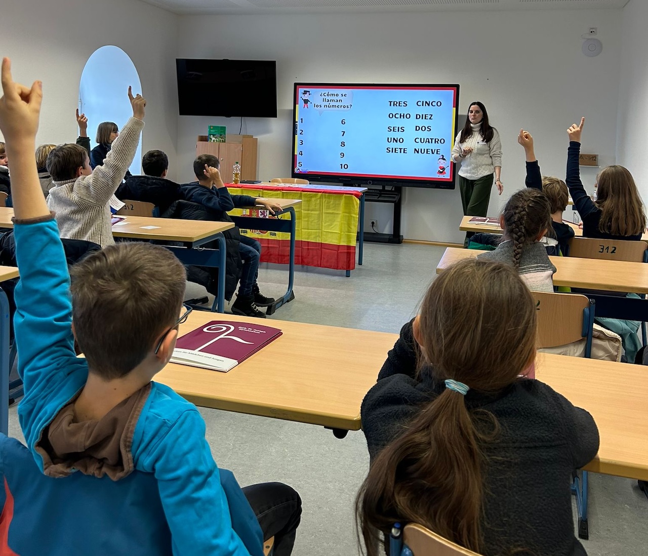 Lehrerin vor digitaler Tafel, darauf spanische Zahlen, Schüler:innen blicken in ihre Richtung