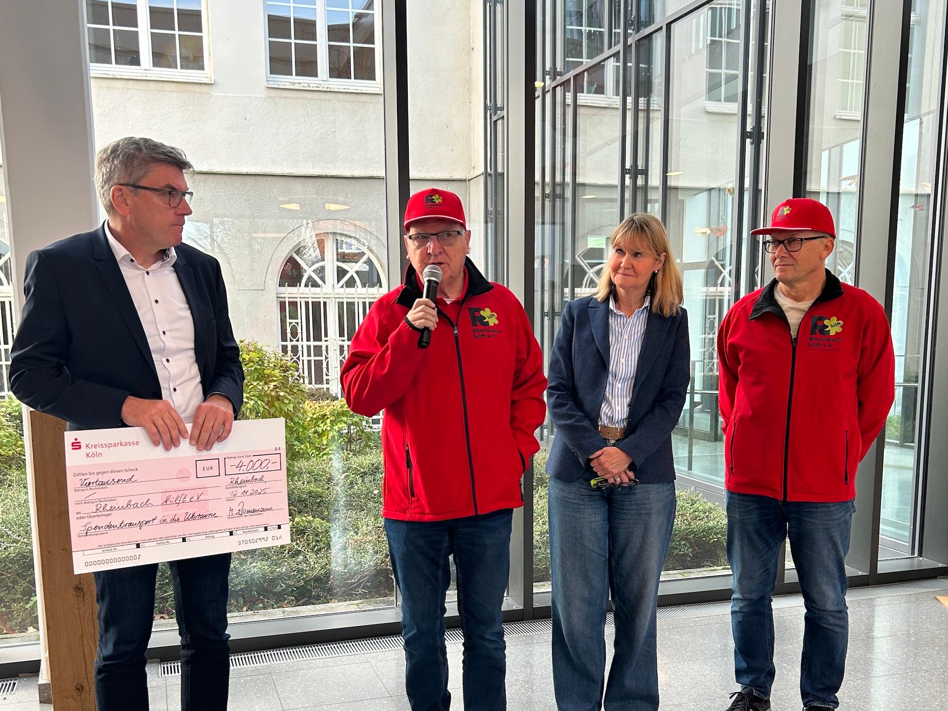 Mann hält großen Scheck in der Hand, daneben zwei Männer in roten Jacken mit roten Mützen, einer spricht in ein Mikro, zwischen den beiden eine Frau