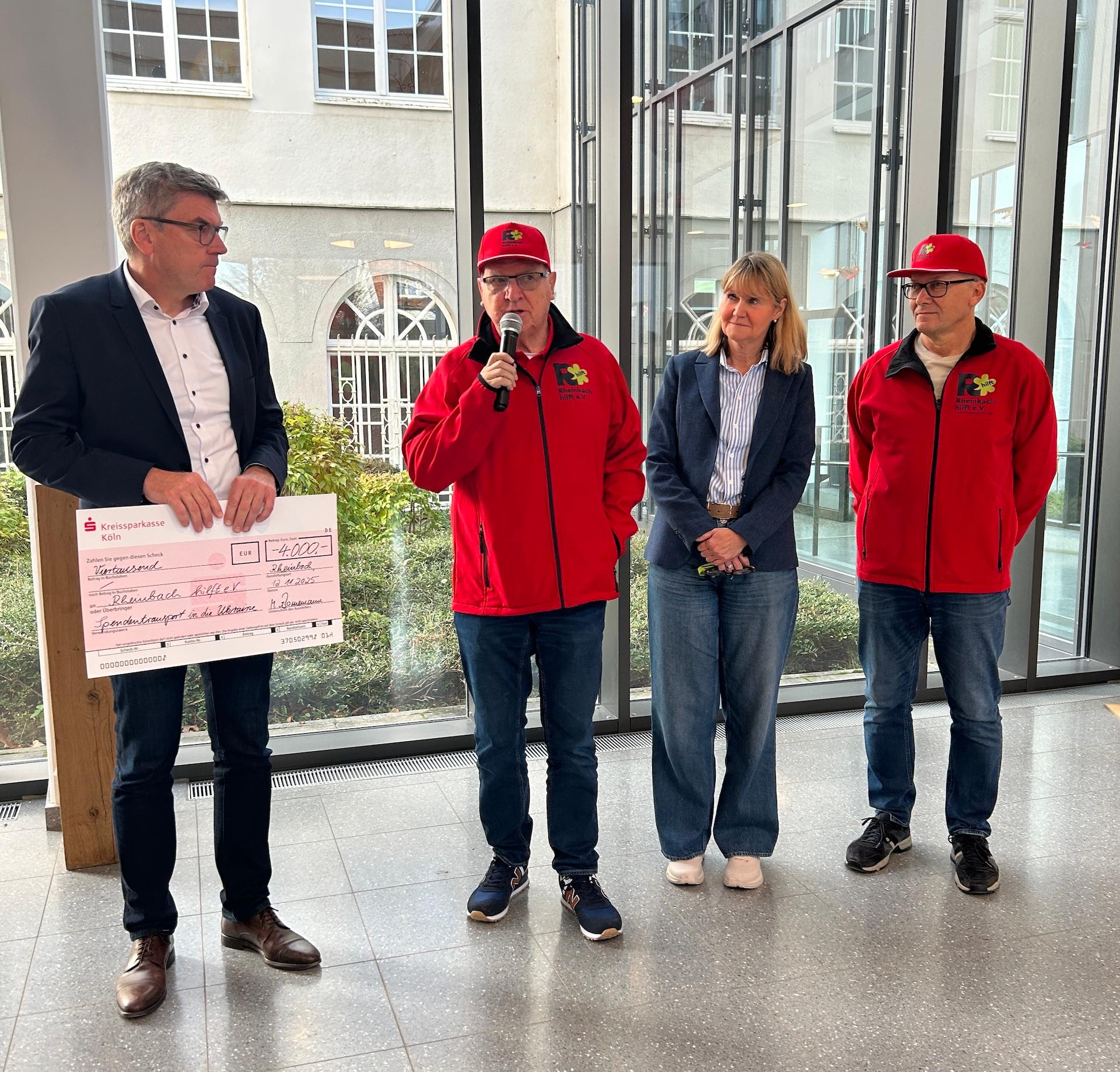 Mann hält großen Scheck in der Hand, daneben zwei Männer in roten Jacken mit roten Mützen, einer spricht in ein Mikro, zwischen den beiden eine Frau