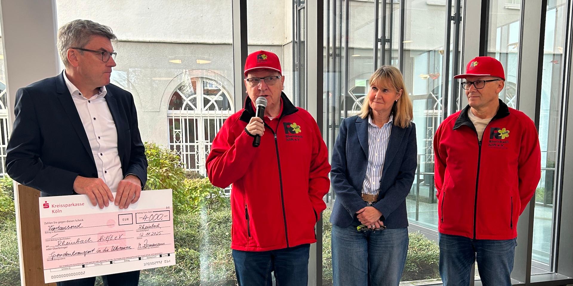 Mann hält großen Scheck in der Hand, daneben zwei Männer in roten Jacken mit roten Mützen, einer spricht in ein Mikro, zwischen den beiden eine Frau