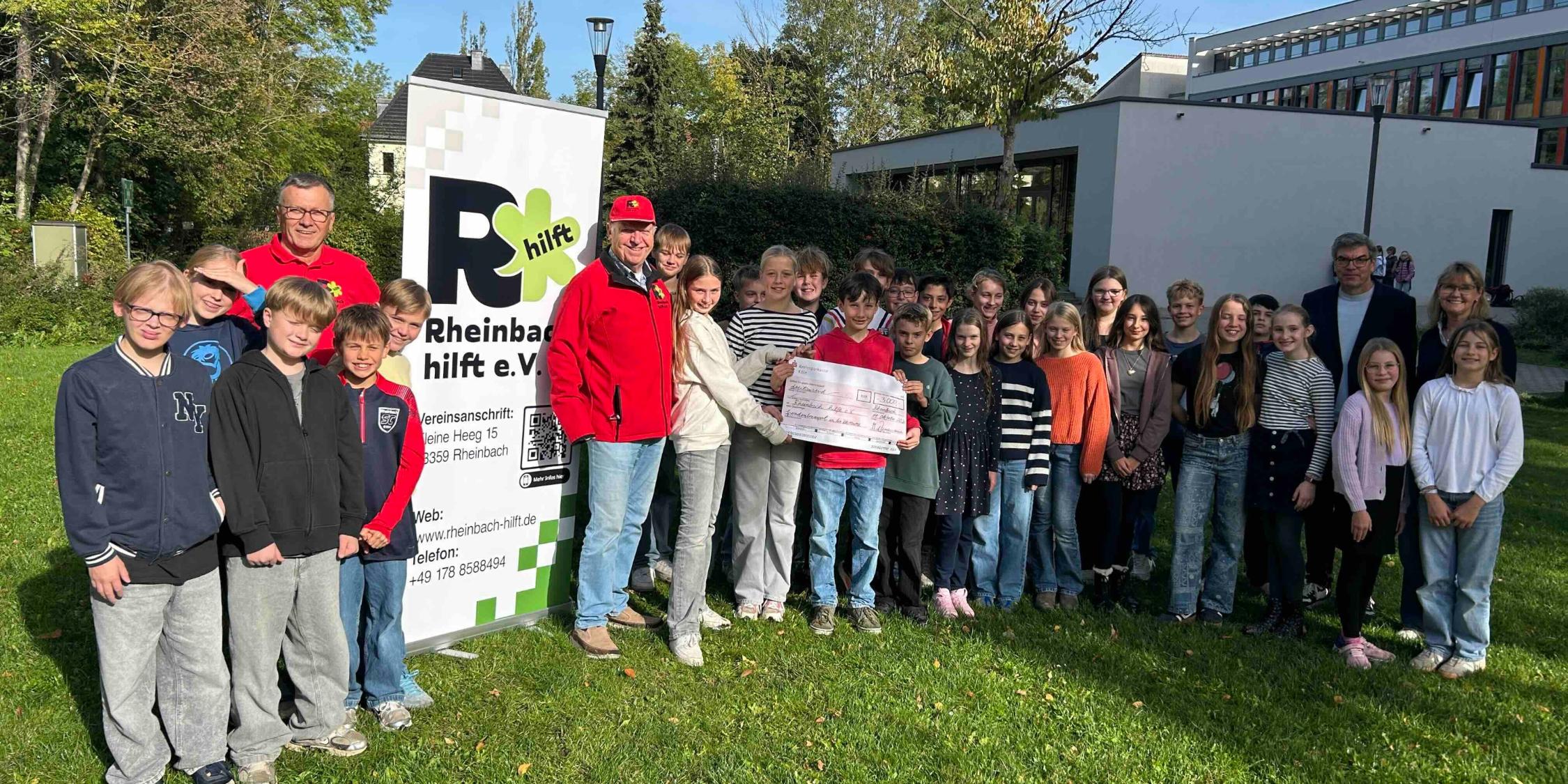Ein Spendenscheck vom Schulfest an 'Rheinbach hilft' Schulklasse, ein Junge hält einen Scheck, zwei Männern neben einem Roll-Up mit 'Rheibach hilft', Schulleitung rechts