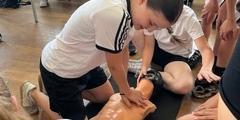 Schülerin übt an einer Puppe Herzdruckmassage, andere Schüler schauen zu