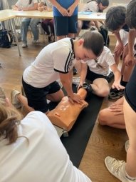 Schülerin übt an einer Puppe Herzdruckmassage, andere Schüler schauen zu