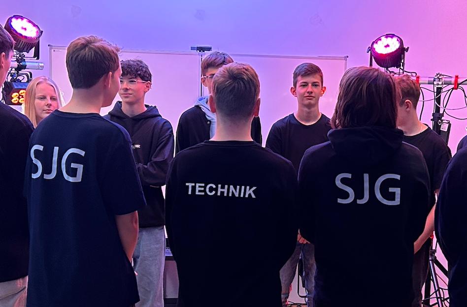 Schülerinnen und Schüler in Hoodys mit Schriftzug TECHNIK im Klassenraum, von Schweinwerfern beleuchtet
