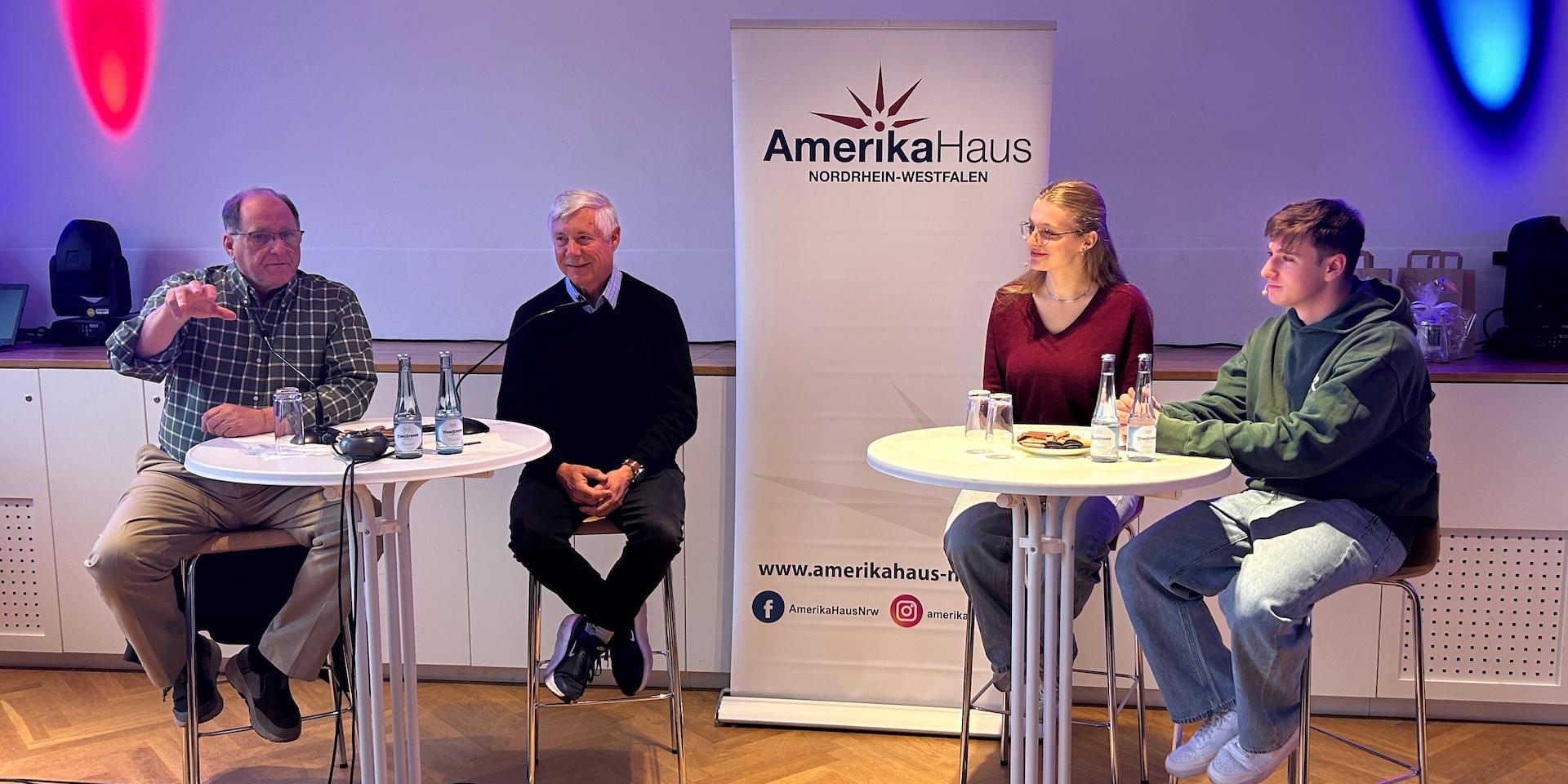 zwei Männer sitzen links auf Barhockern, eine Schülerin, ein Schüler rechts auf Barhockern, dahinter ein Rollup vom Amerika-Haus NRW, rote und blaue Strahler