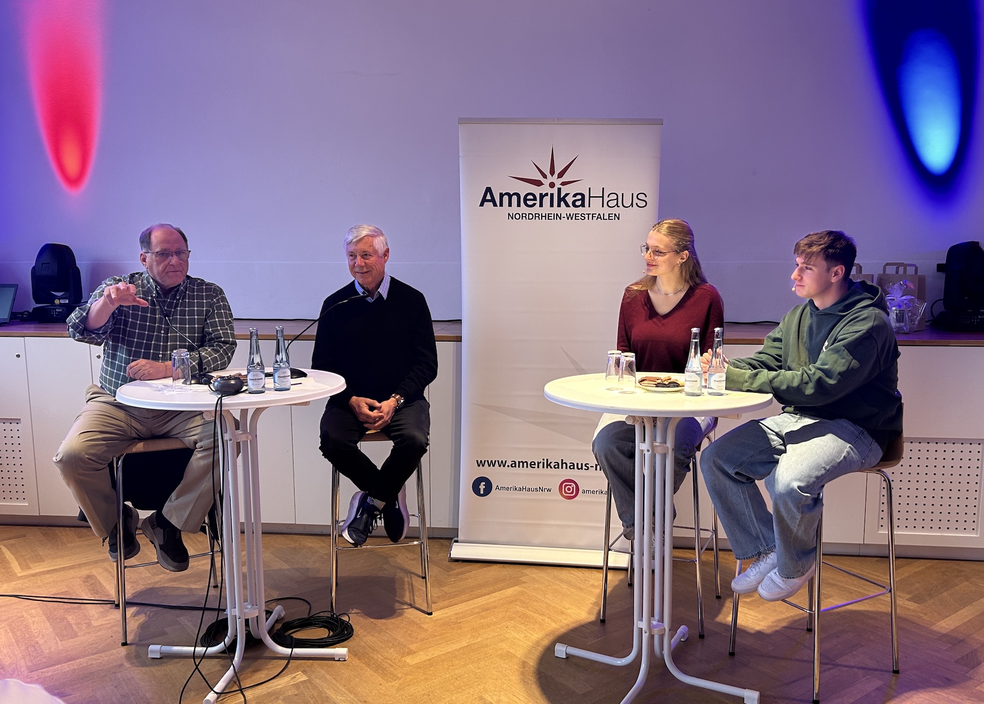 zwei Männer sitzen links auf Barhockern, eine Schülerin, ein Schüler rechts auf Barhockern, dahinter ein Rollup vom Amerika-Haus NRW, rote und blaue Strahler