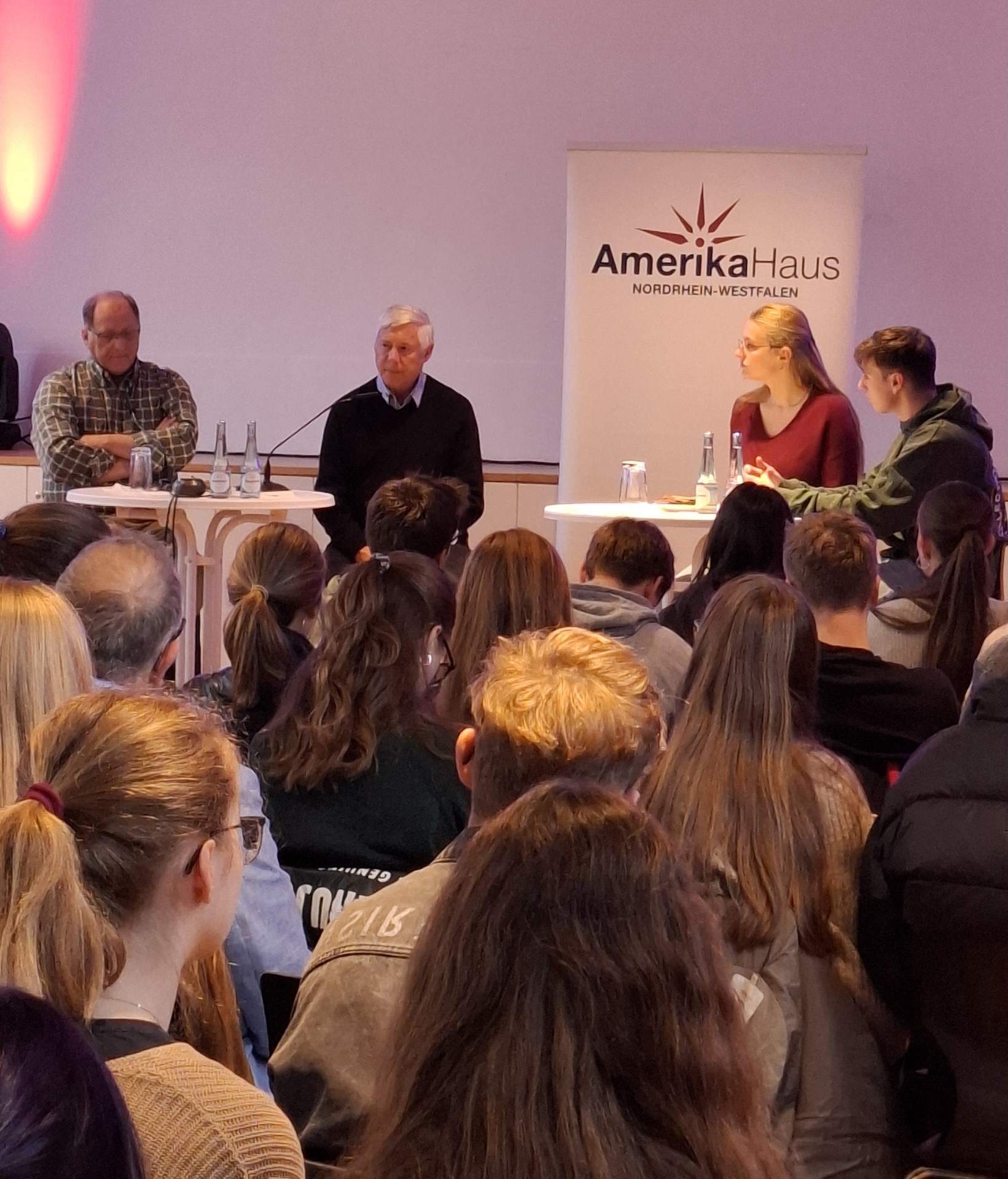 Schülergruppe sitzend von hinten, blicken auf zwei Männer, eine Schülerin und deinen Schüler auf Barhockern