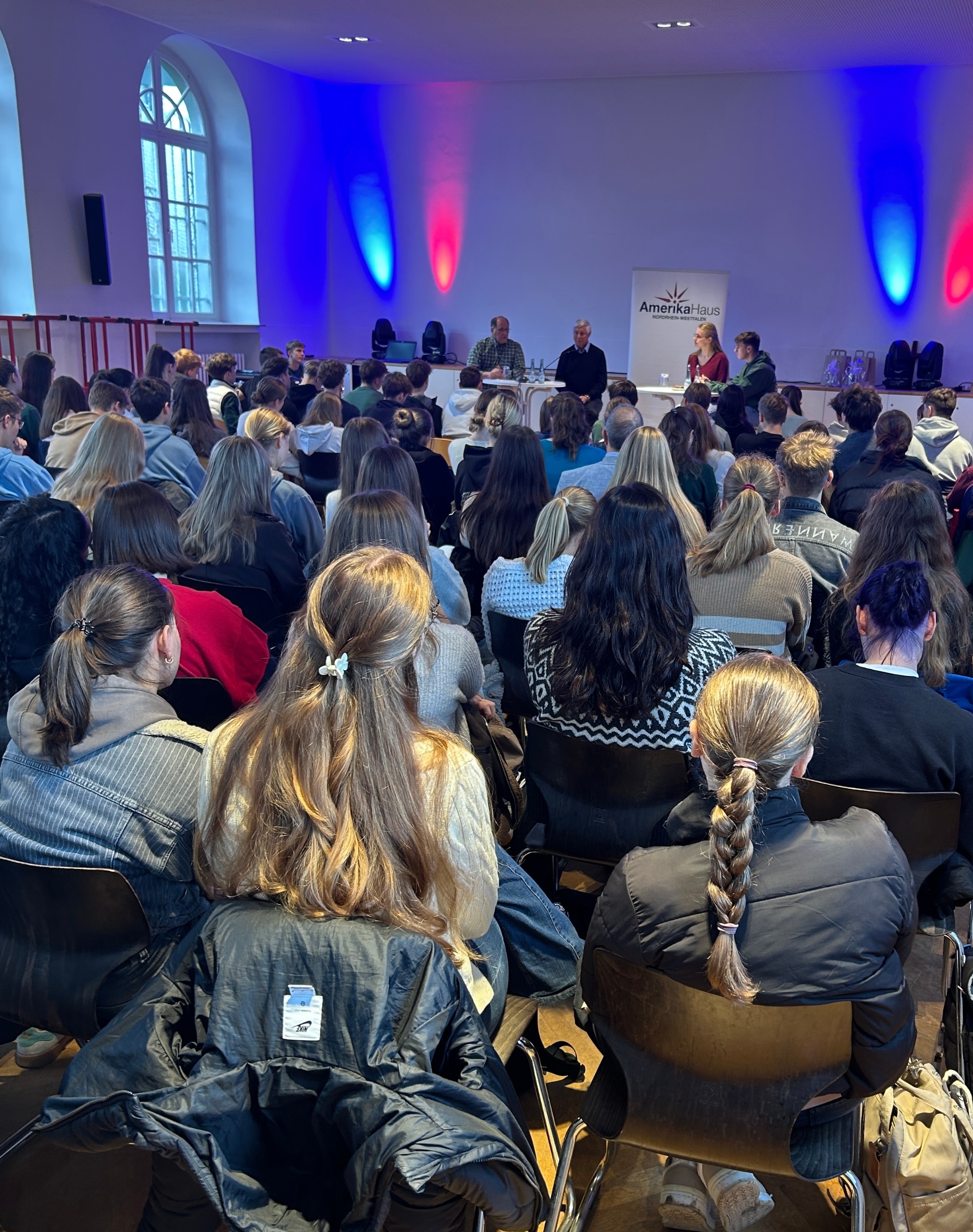 Schüler:innen sitzen in Reihen im Saal, blicken nach vorn zu vier Diskutierenden