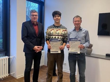 Drei Männer (Schulleiter, Schüler, Lehrer) stehen nebeneinander, Schüler und Lehrer halten Urkunden