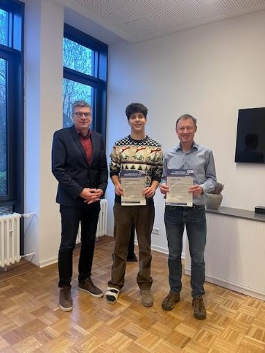 Drei Männer (Schulleiter, Schüler, Lehrer) stehen nebeneinander, Schüler und Lehrer halten Urkunden