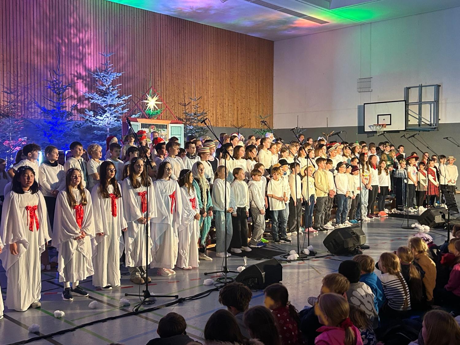 Kinder als Engel verkleidet, daneben weitere Kinder im Chor, davor sitzend Kinder im Publikum