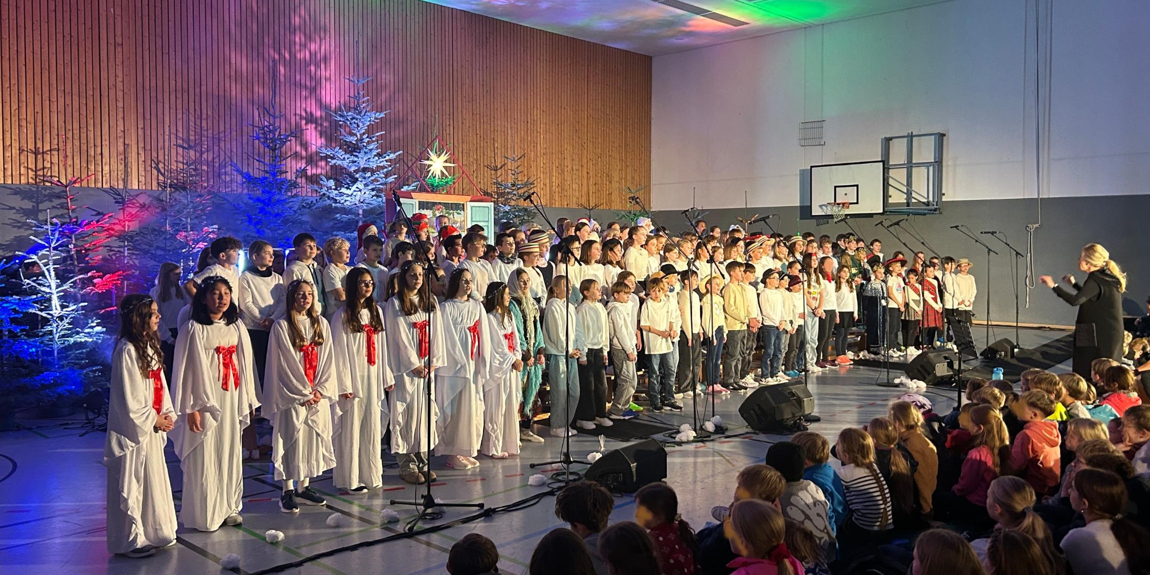 Kinder als Engel verkleidet, daneben weitere Kinder im Chor, davor sitzend Kinder im Publikum