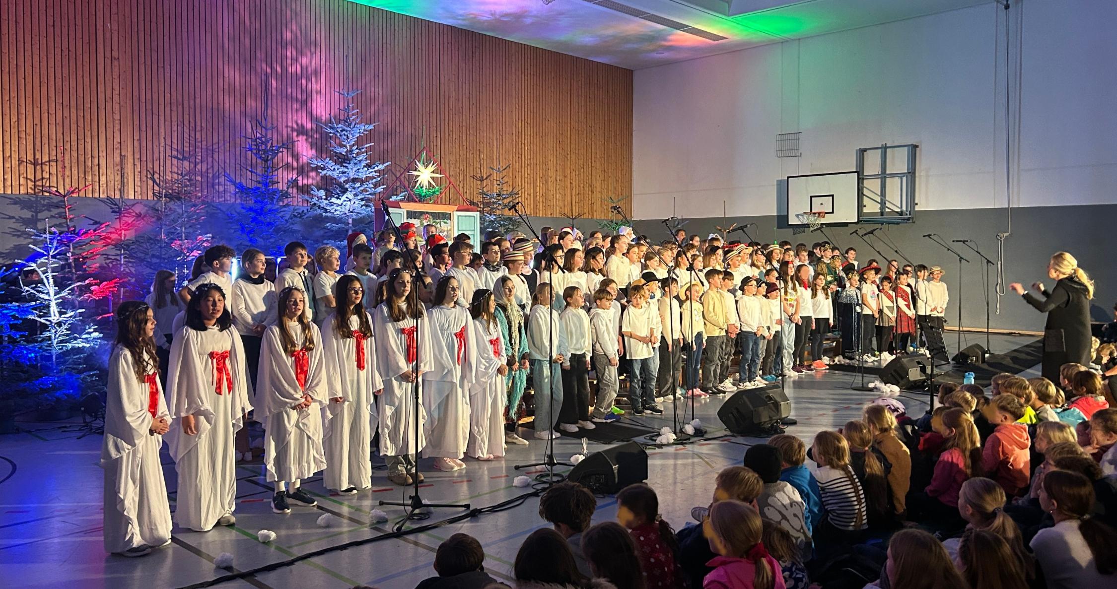 Kinder als Engel verkleidet, daneben weitere Kinder im Chor, davor sitzend Kinder im Publikum