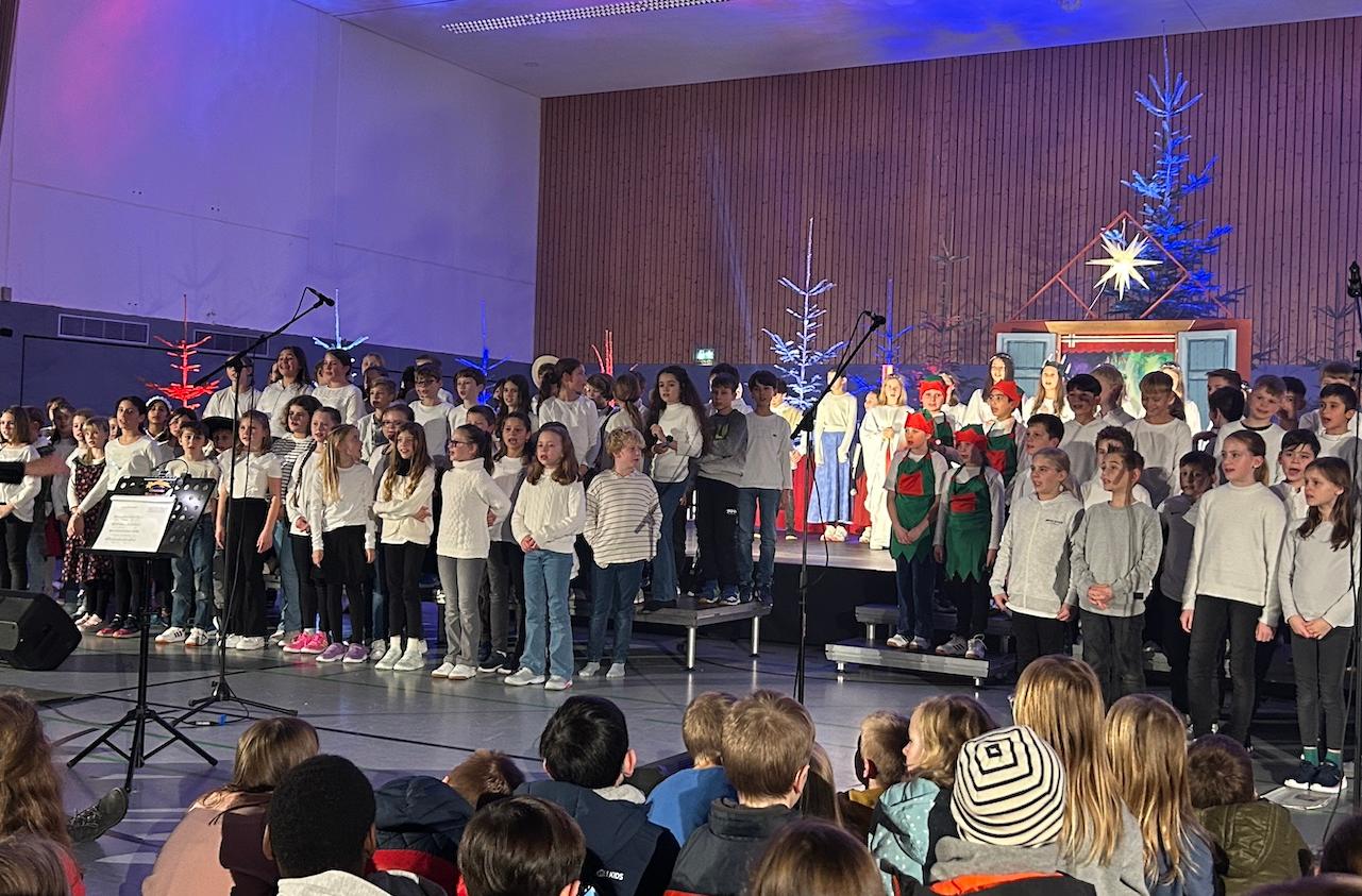 große Gruppe Kinder auf der Bühne, rechts zwei Mädchen in ungarischer Tracht, im Hintergrund Tannenbäume, vorne sitzende Kinder auf dem Fußboden, vor den Kindern Dirigentin