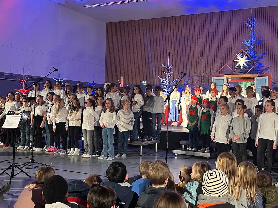 große Gruppe Kinder auf der Bühne, rechts zwei Mädchen in ungarischer Tracht, im Hintergrund Tannenbäume, vorne sitzende Kinder auf dem Fußboden, vor den Kindern Dirigentin