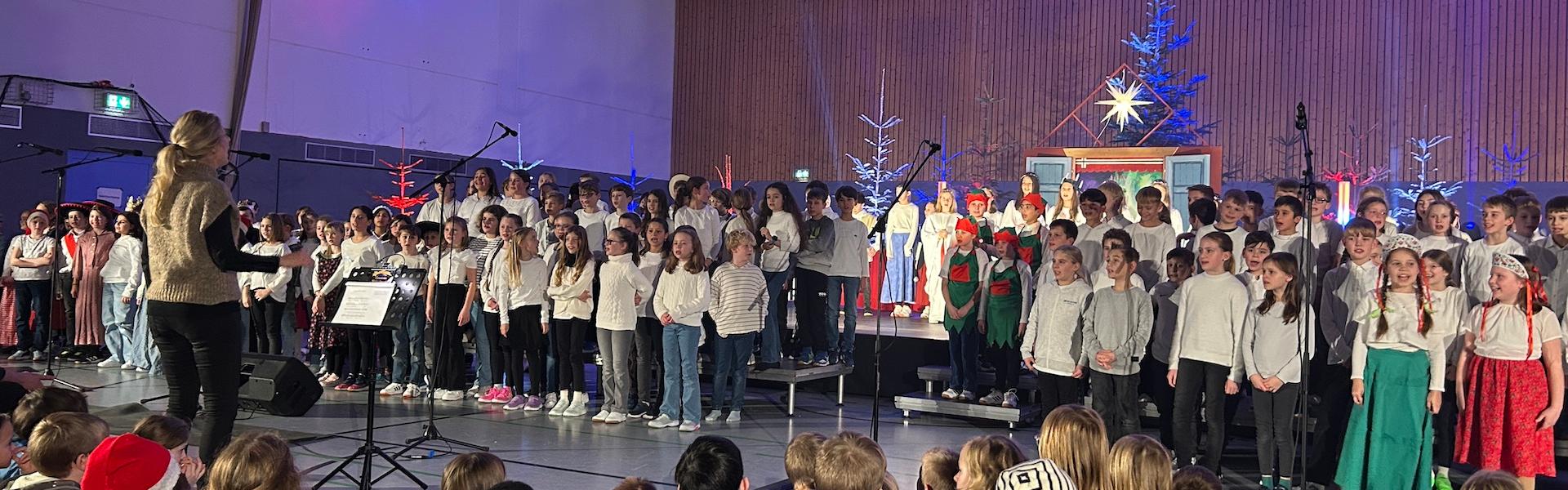 große Gruppe Kinder auf der Bühne, rechts zwei Mädchen in ungarischer Tracht, im Hintergrund Tannenbäume, vorne sitzende Kinder auf dem Fußboden, vor den Kindern Dirigentin