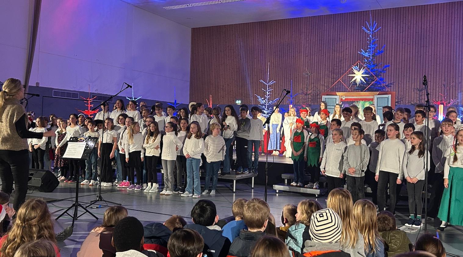 große Gruppe Kinder auf der Bühne, rechts zwei Mädchen in ungarischer Tracht, im Hintergrund Tannenbäume, vorne sitzende Kinder auf dem Fußboden, vor den Kindern Dirigentin