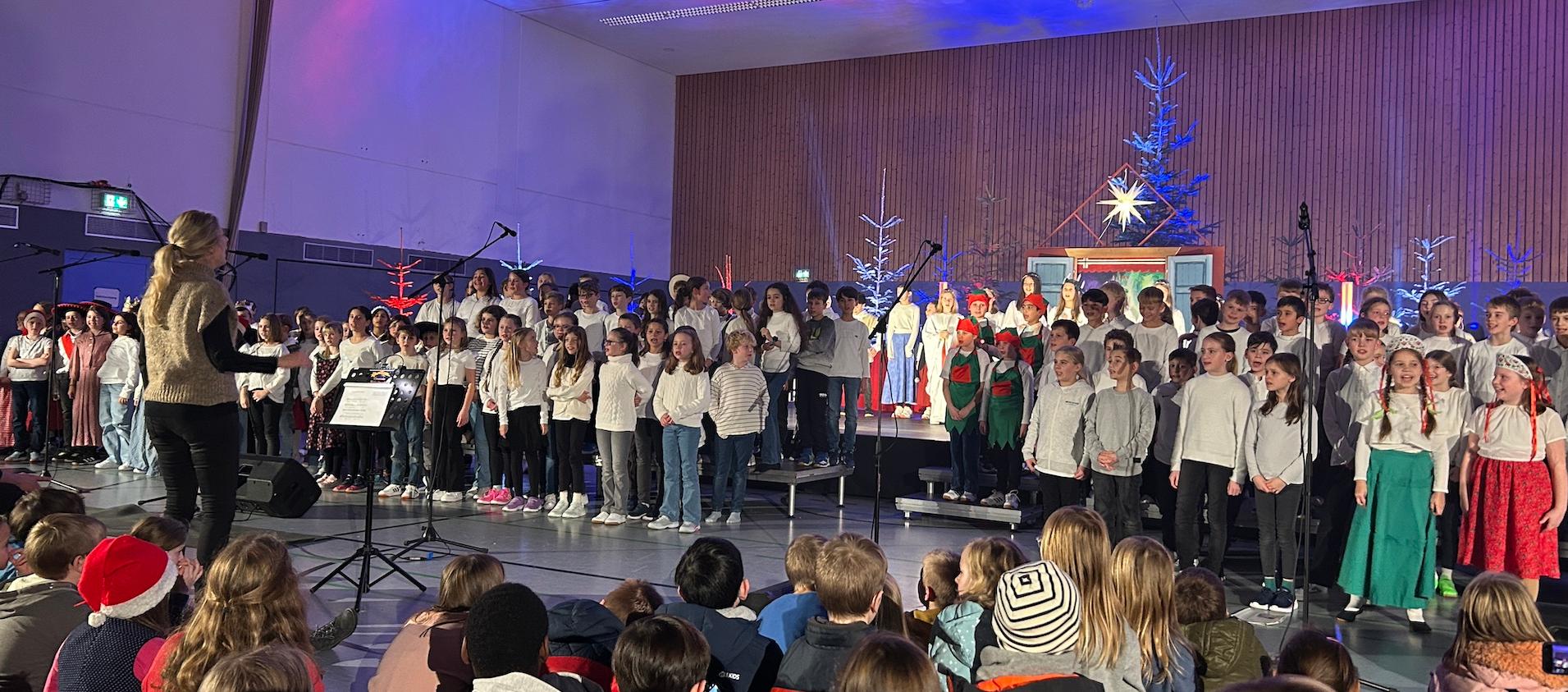 große Gruppe Kinder auf der Bühne, rechts zwei Mädchen in ungarischer Tracht, im Hintergrund Tannenbäume, vorne sitzende Kinder auf dem Fußboden, vor den Kindern Dirigentin