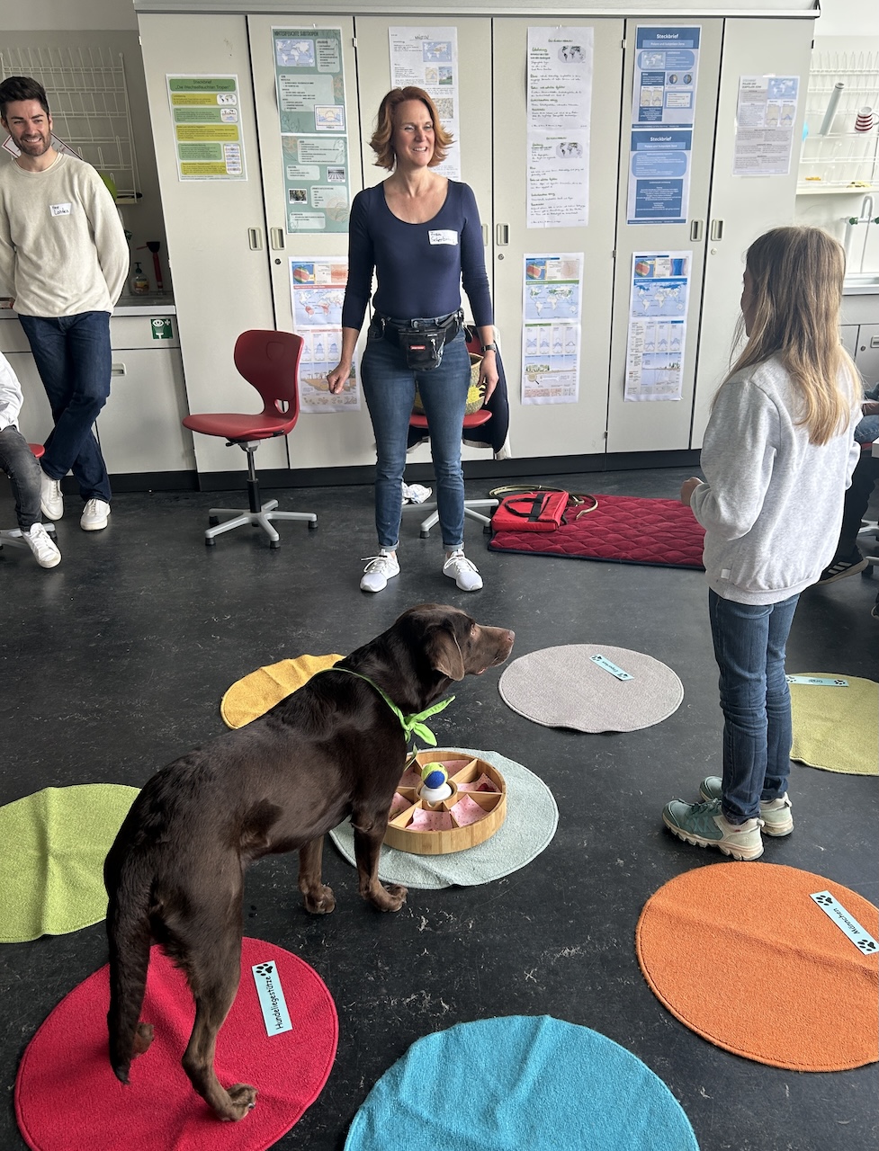 Schulhund blickt zu Schülerin, im Hintergrund Lehrerin und Lehrer
