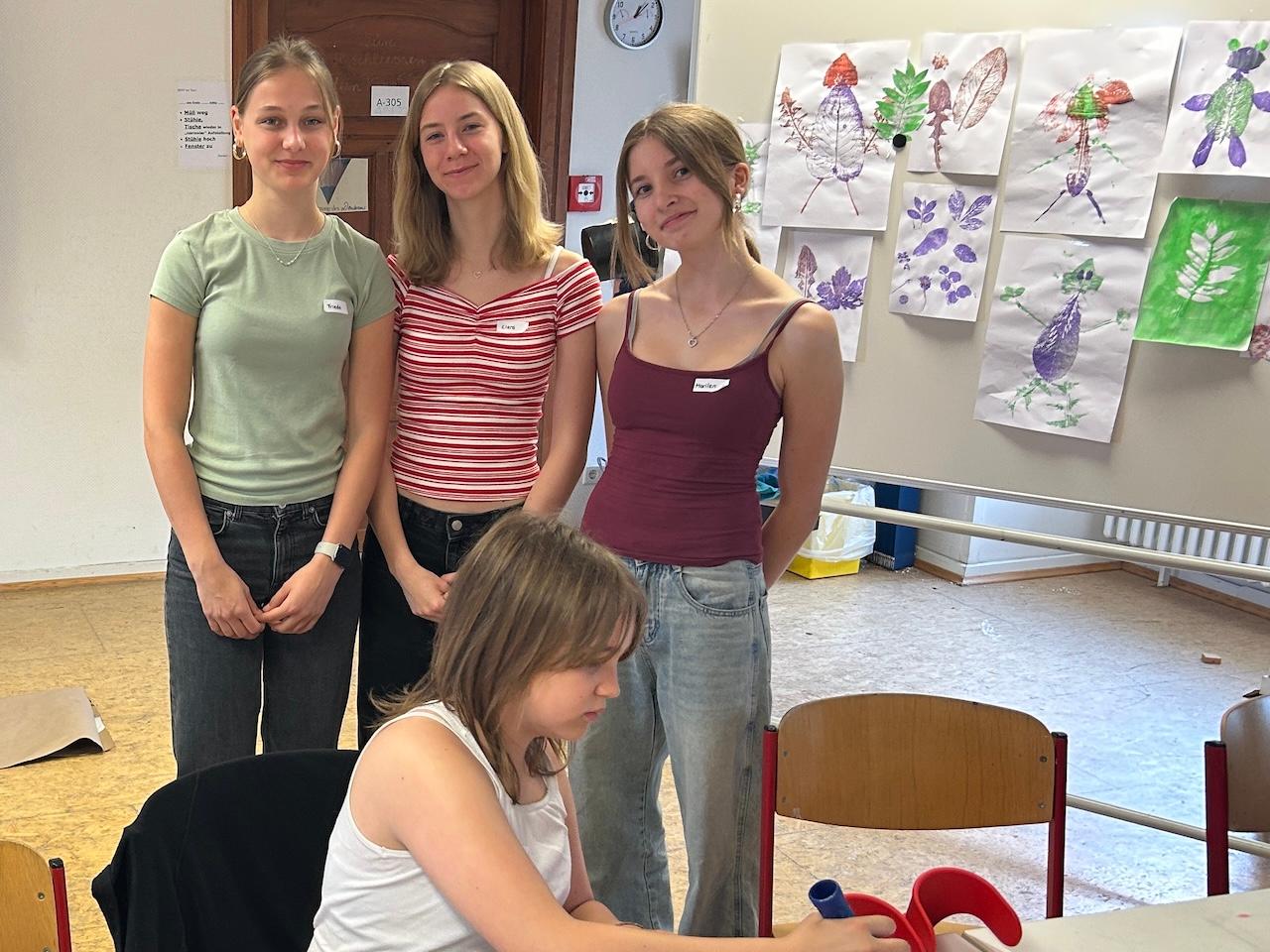 3 Schülerinnen vor einer Pinnwand mit gedruckten Bildern, vorne eine Schülerin mit Farbrolle