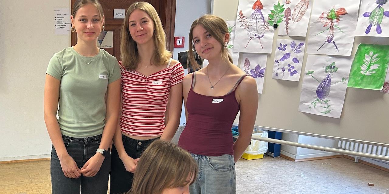 3 Schülerinnen vor einer Pinnwand mit gedruckten Bildern, vorne eine Schülerin mit Farbrolle