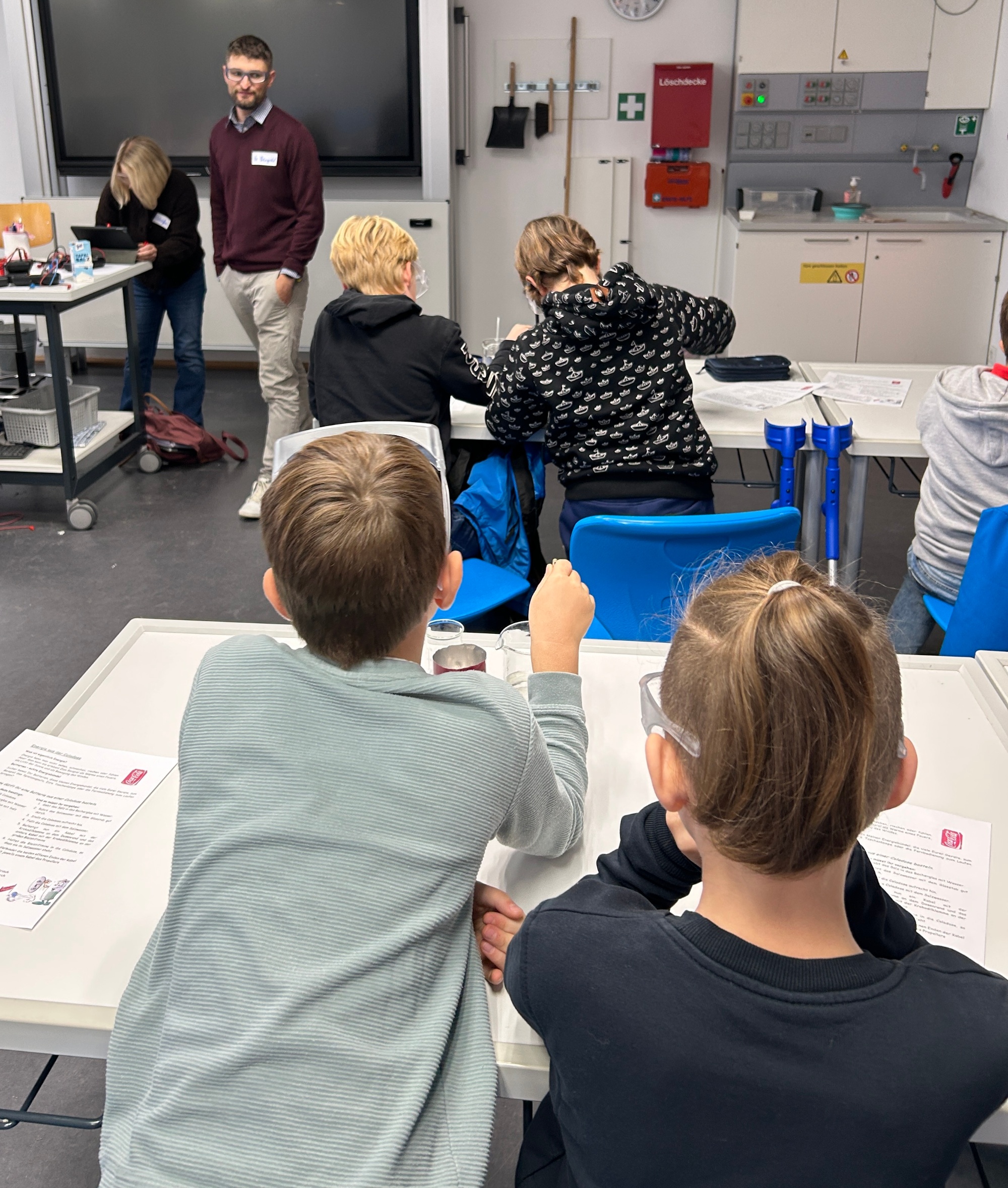 Schüler an Tischen im Chemieraum, Lehrerin und Lehrer vorn