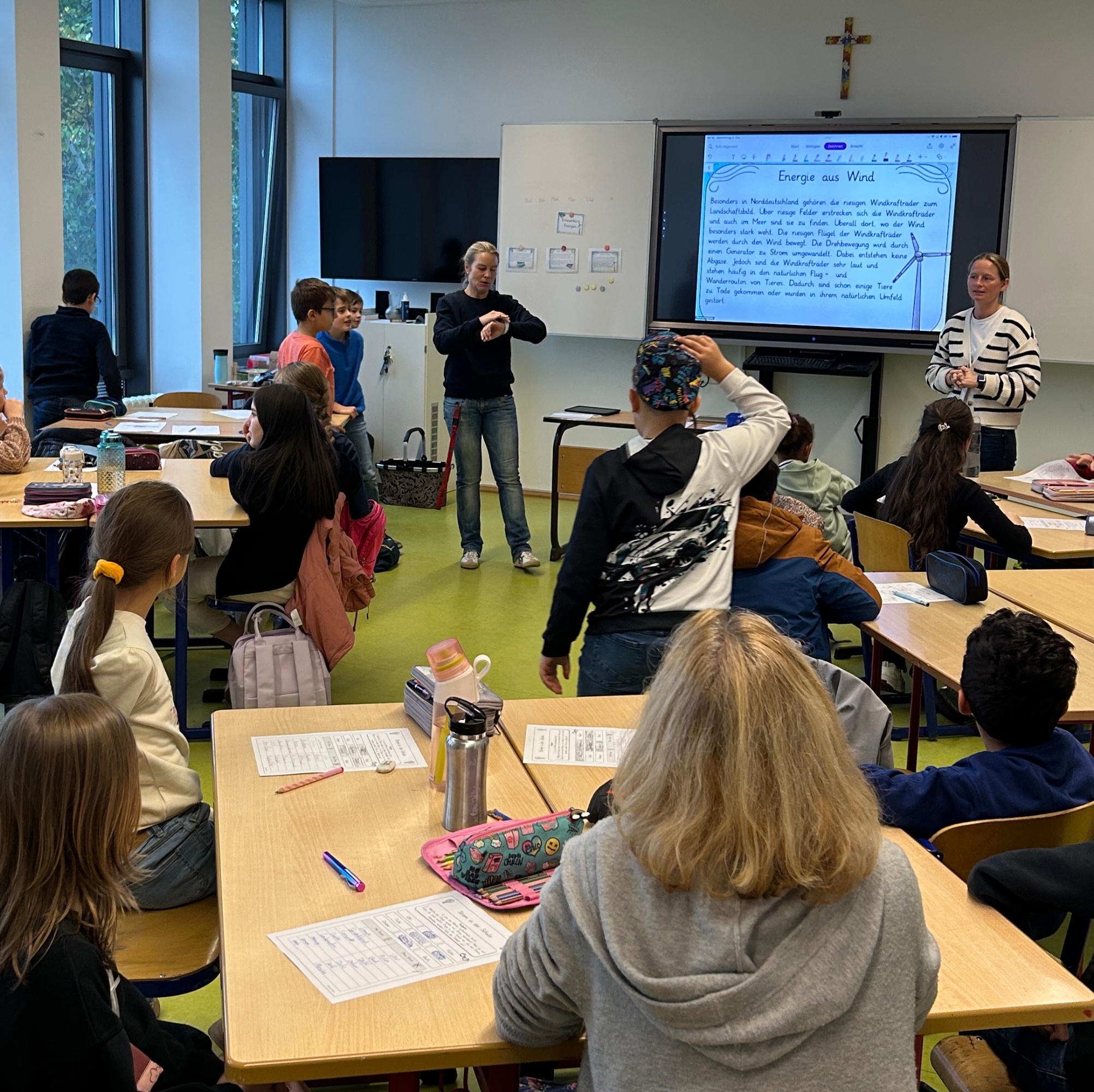 Schülerinnen und Schüler an Gruppentischen, zwei Lehrerinnen vor einer digitalen Tafel